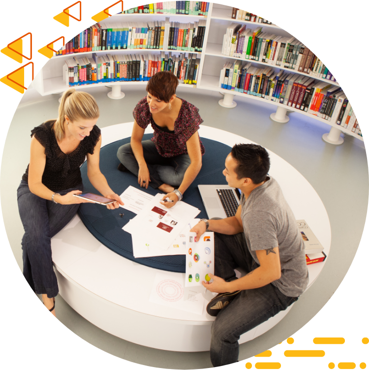 Aerial photo of three students collaborating on design work while seated on a large ottoman, books are in the background.